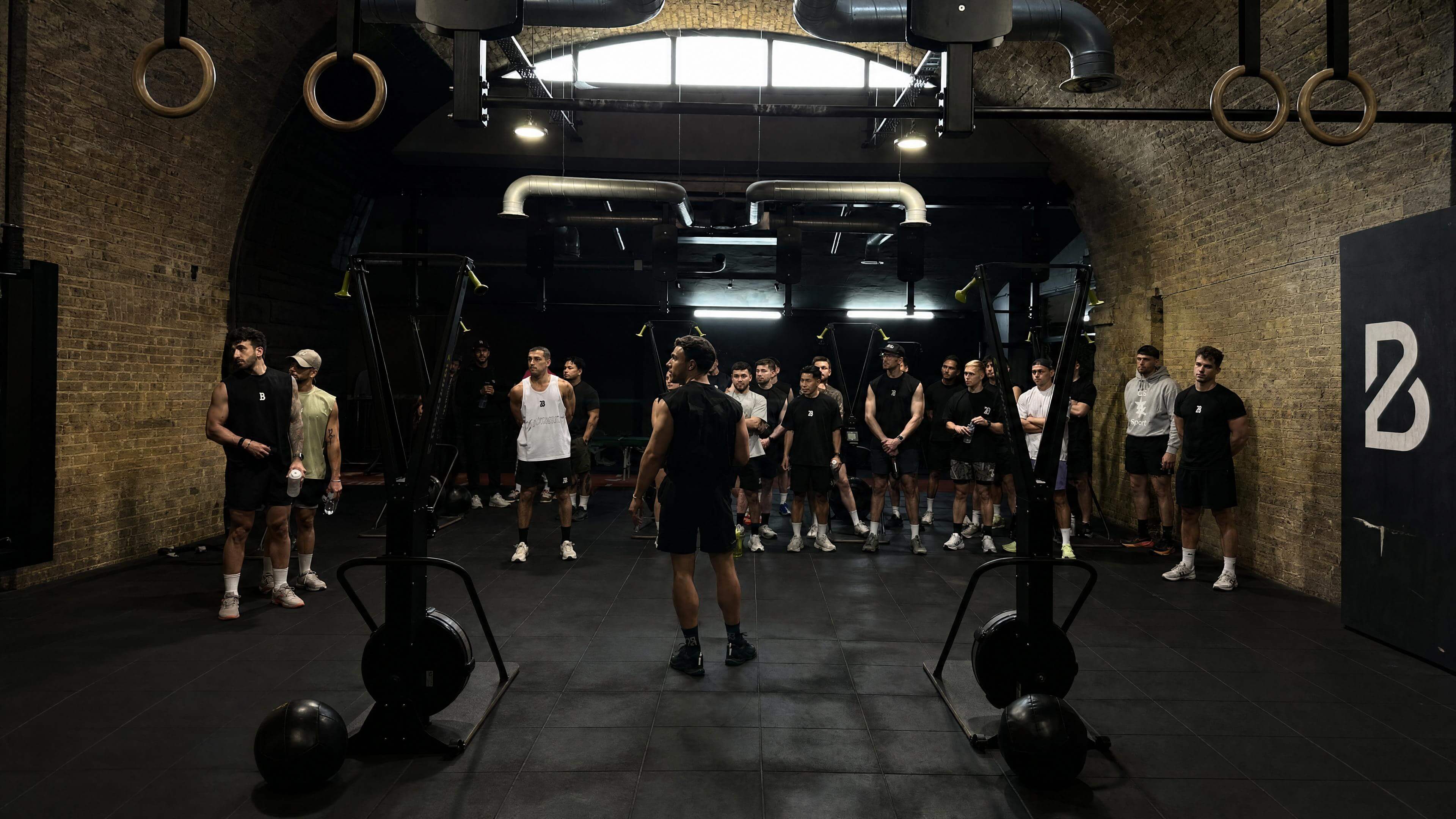 Black and white photograph — Teji coaching a full class of men in a brick-arched gym