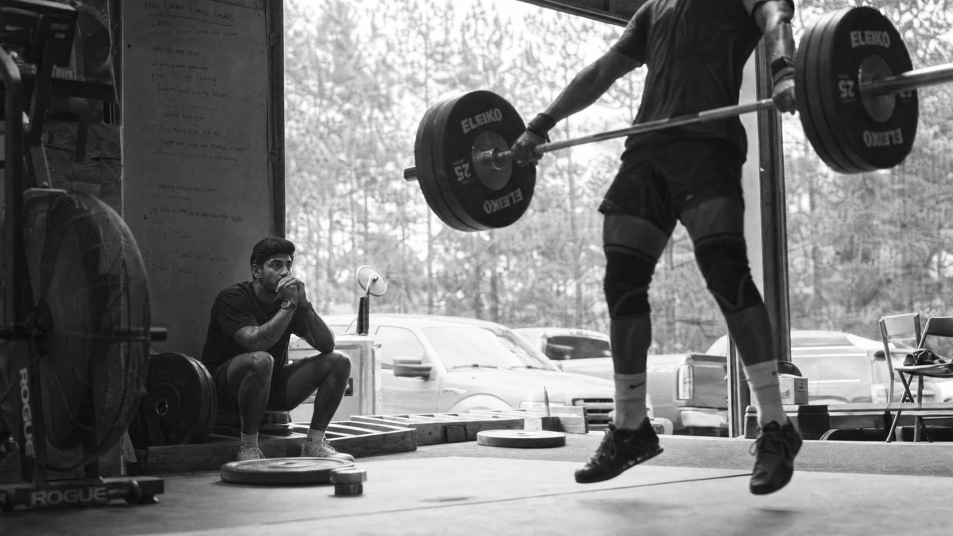Black and white photograph — Teji seated at the edge of an outdoor gym, watching an athlete catch a loaded barbell mid-lift